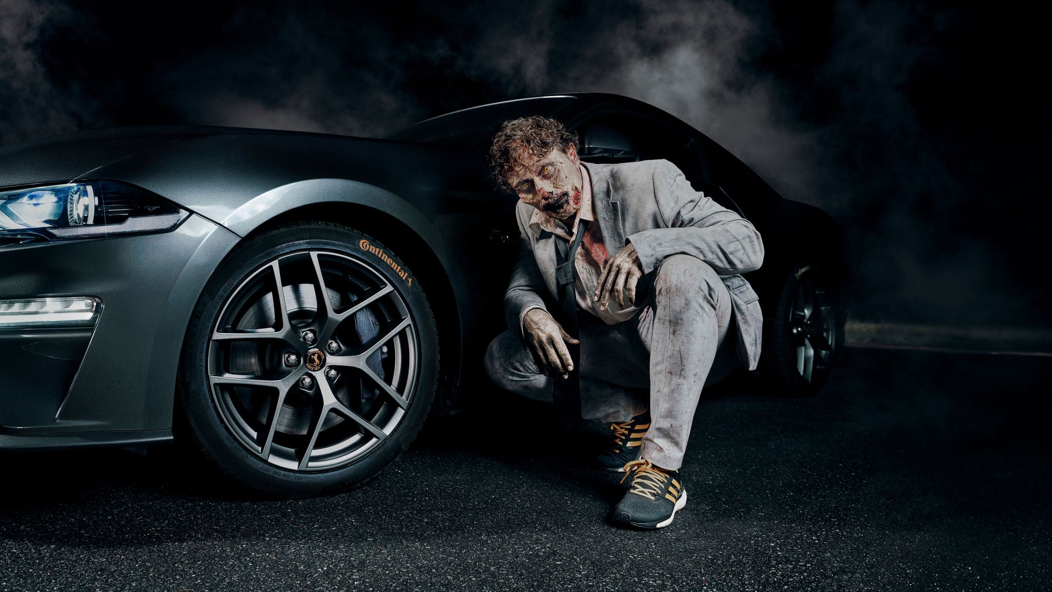 A zombie, wearing a grey suit, is crouching next to the Continental front tire.