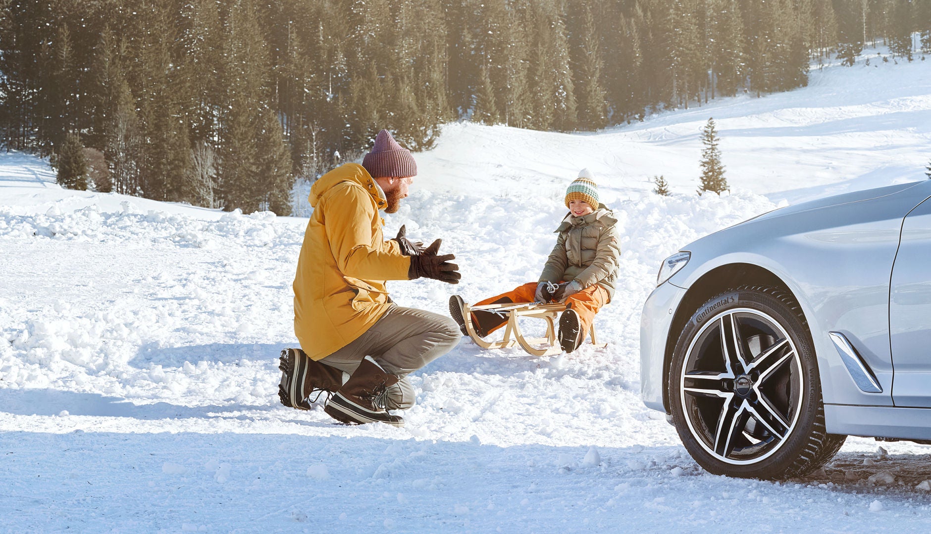 Un enfant est assis sur une luge, tandis qu'un adulte vêtu d'une veste jaune est assis à proximité dans un paysage enneigé. Au premier plan, une voiture argentée est garée.