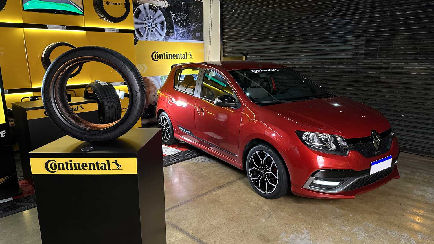 (La imagen muestra un espacio de exposición. A la derecha, el automóvil Renault Sandero, que presenta un diseño deportivo, en color rojo, con detalles en negro, las ruedas con neumáticos ExtremeContact Sport 02. El automóvil se encuentra estacionado sobre un concreto pulido. A la izquierda, se muestra una exposición de llantas Continental, con un destaque para la llanta ExtremeContact Sport 02 sobre una base de pedestal. Al fondo, se ven otros expositores y estanterías con más llantas en exposición, todos con iluminación predominantemente amarilla, que también forma parte de la colección. parte de la identidad visual de la marca. El ambiente del lugar es el de una vitrina de productos automotrices, con foco en el desempeño y la tecnología de las llantas y vehículos)